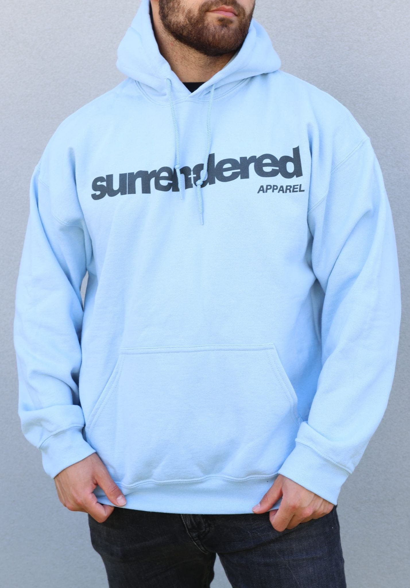 A man wearing a Surrendered Apparel Heavyblend Hoodie in Light Blue. The Surrendered Apparel Logo in black sits across the chest of the Hoodie. The hoodie has a loose and comfortable fit 