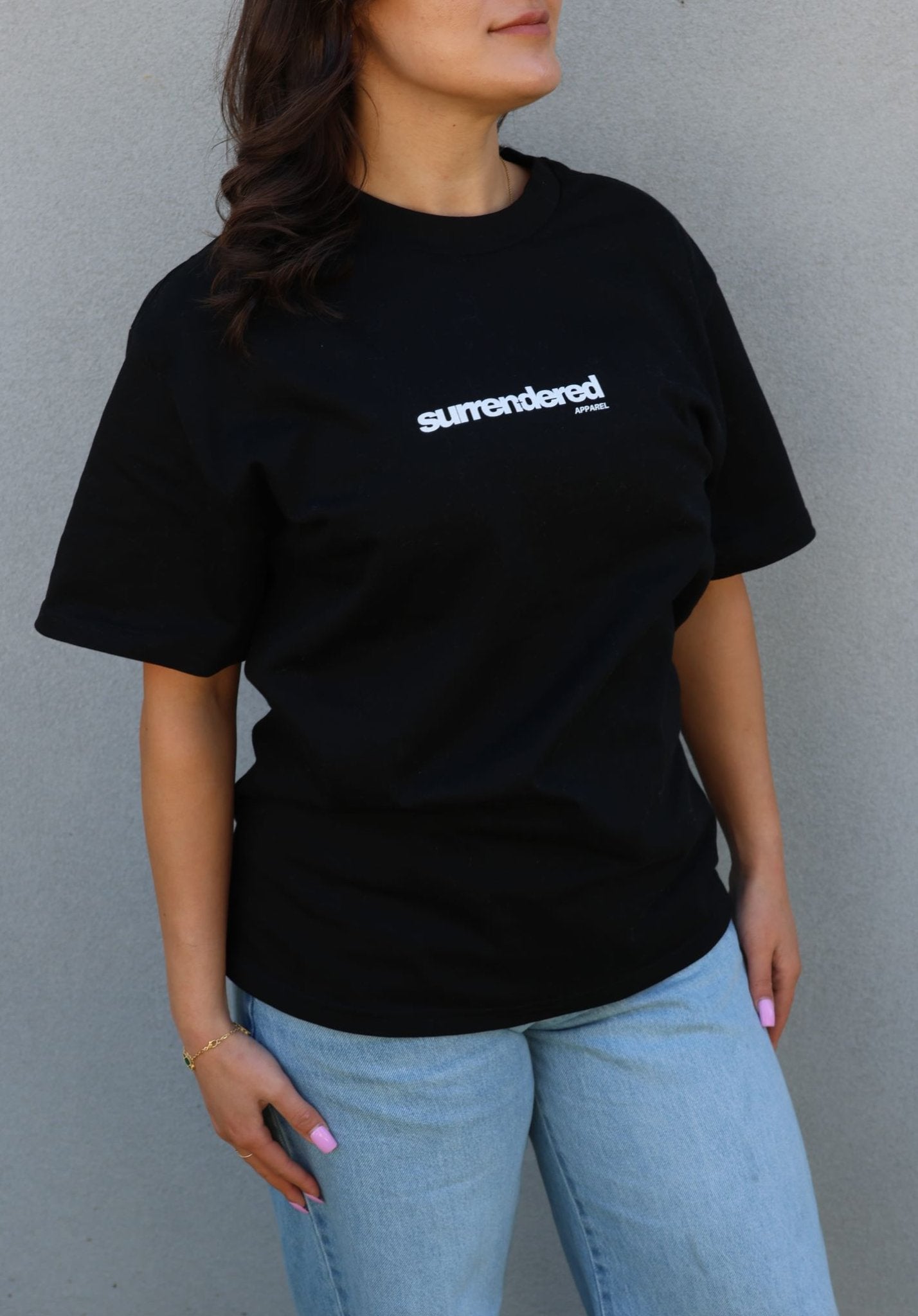 Woman wearing a Surrendered Apparel Unisex Oversize T-shirt in Black. Shirt is thick cotton in black colour, white Surrendered Apparel Logo across the front of the chest. Woman is wearing blue jeans