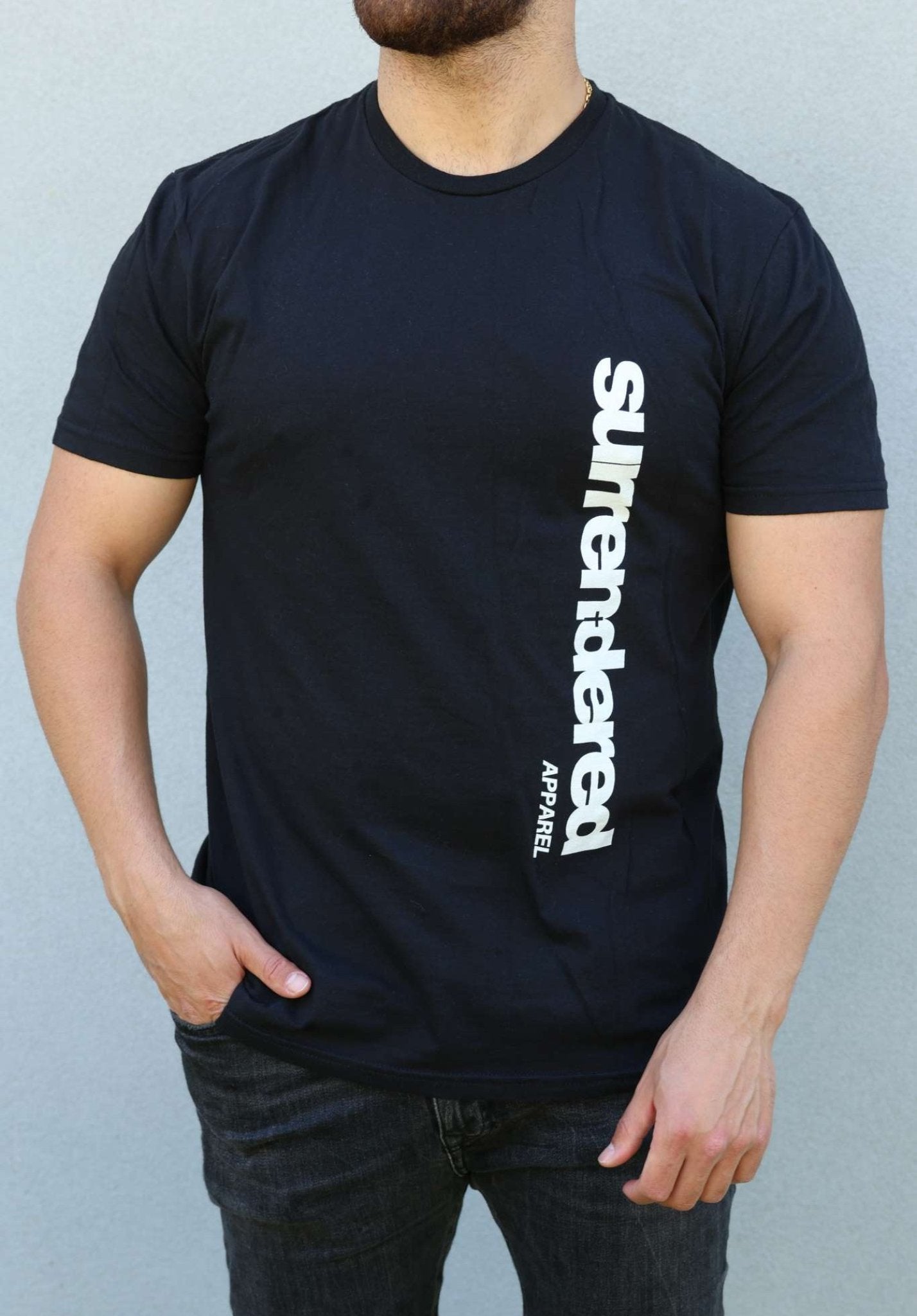 Man wearing a Surrendered Apparel Softstyle T-shirt. Shirt is Navy Blue with Surrendered Apparel Logo in white positioned vertically on the left side of the shirt. The man is tanned and wearing black jeans